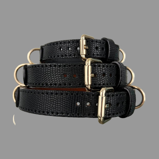 Three black lizard-embossed cowhide dog collars stacked together, with polished gold-tone buckles and D-rings.