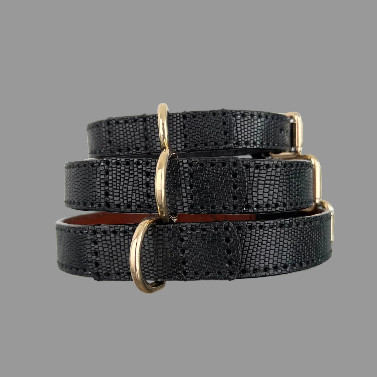 Three stacked black cowhide dog collars showing lizard-scale embossing, black edge stitching and gold-tone D-rings.