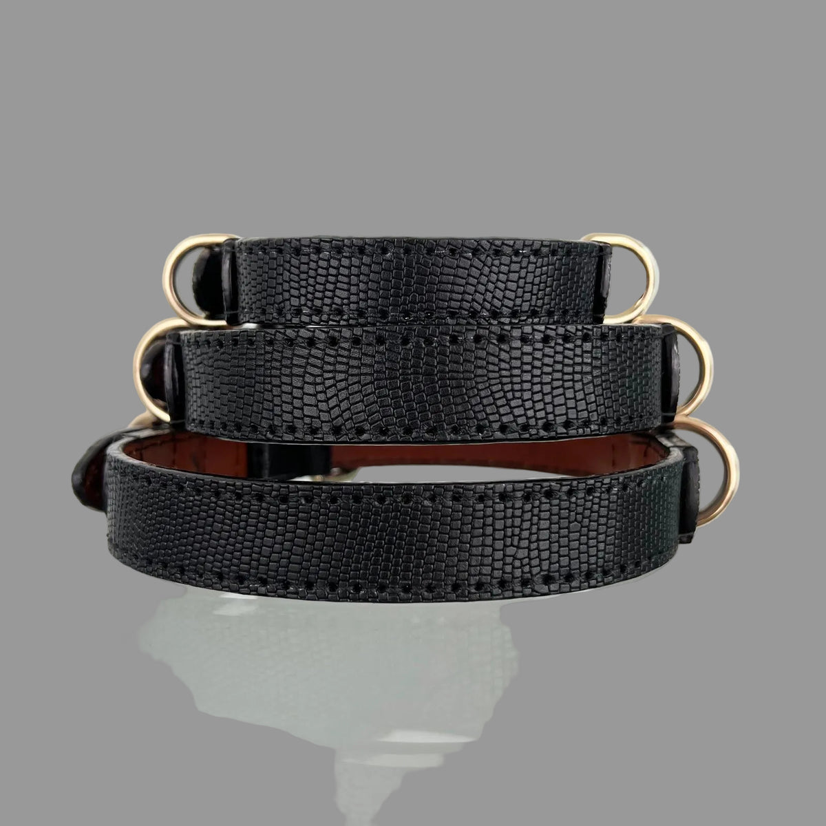 Angled view of three black lizard-embossed leather dog collars with gold-tone buckles, D-rings and matching leather keepers.