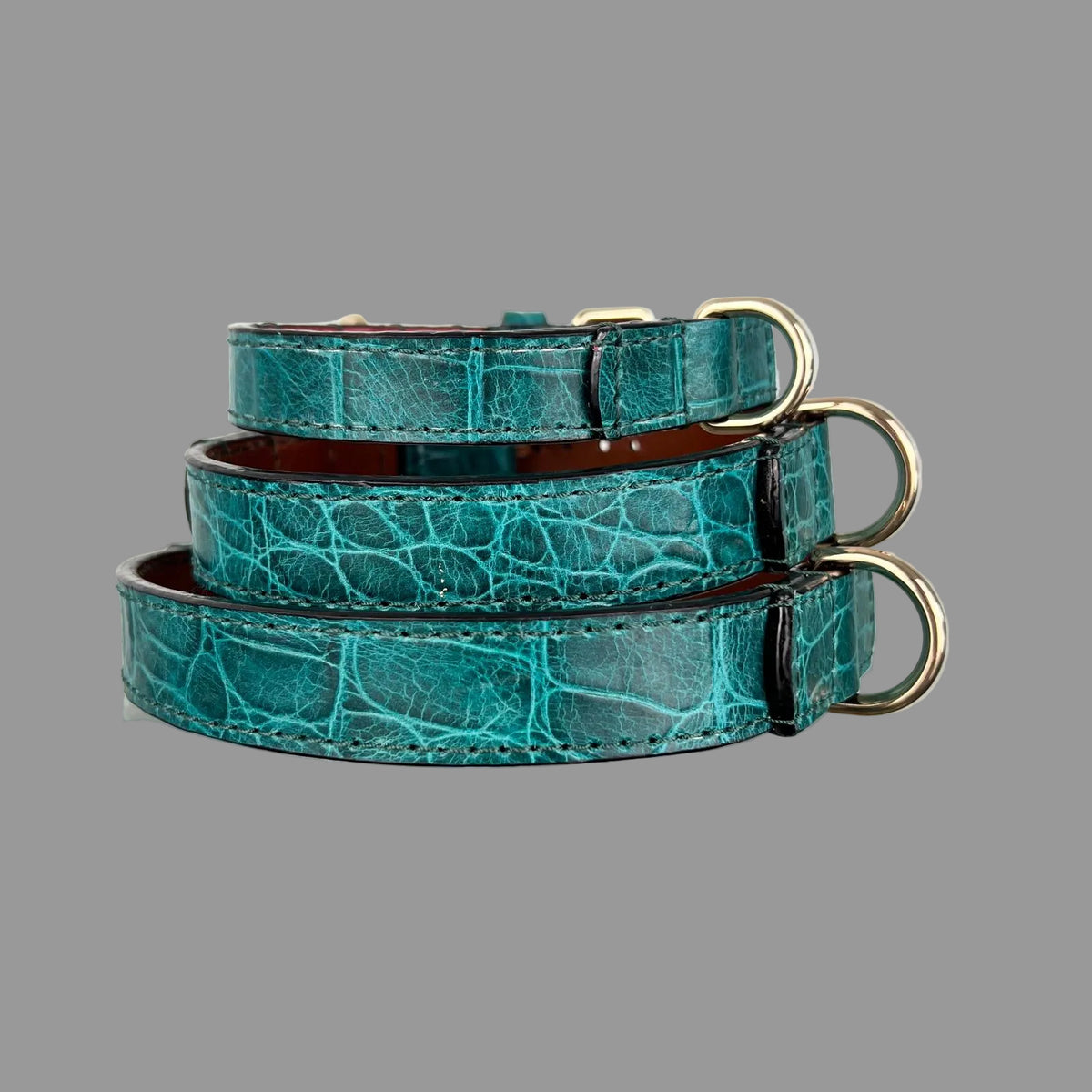 Rear view of three stacked teal leather dog collars showing crocodile-pattern detail and gold-tone D-rings along the right side.