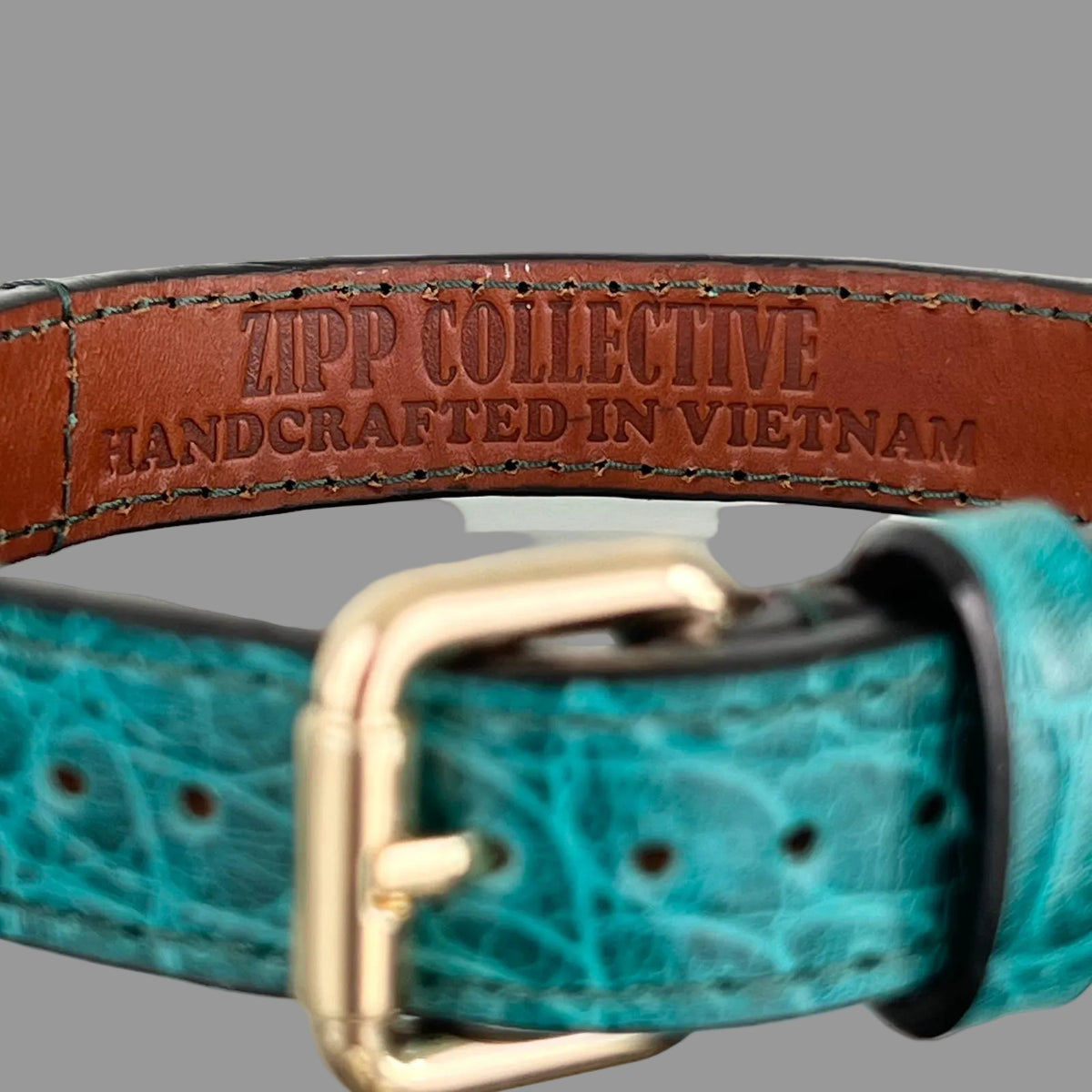 Brown leather lining stamped "ZIPP COLLECTIVE — HANDCRAFTED IN VIETNAM", with the teal embossed strap and gold-tone buckle in front.