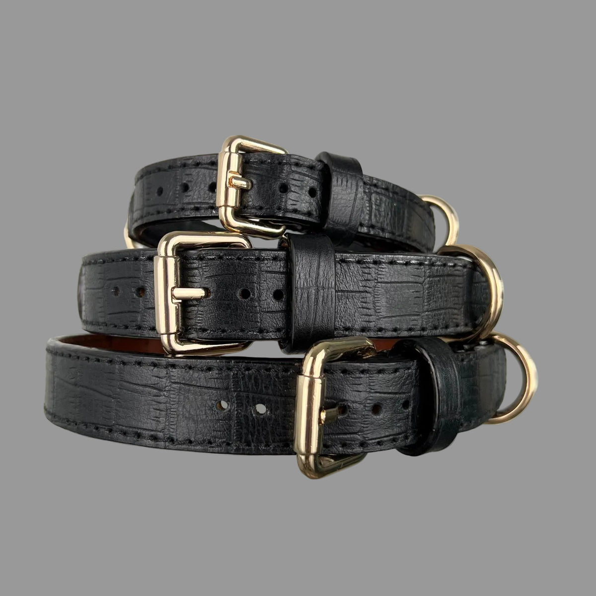 Three black crocodile-embossed leather dog collars stacked to show their widths, gold-tone buckles and black stitching.