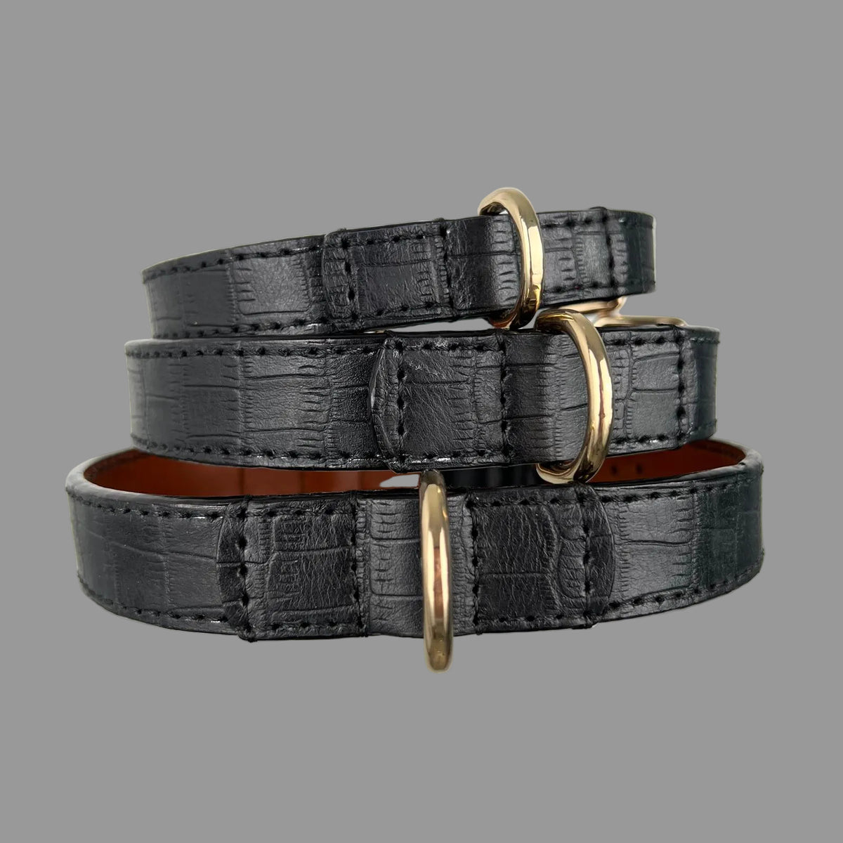 Side view of stacked black crocodile-embossed dog collars showing gold-tone D-rings and stitched leather reinforcement.