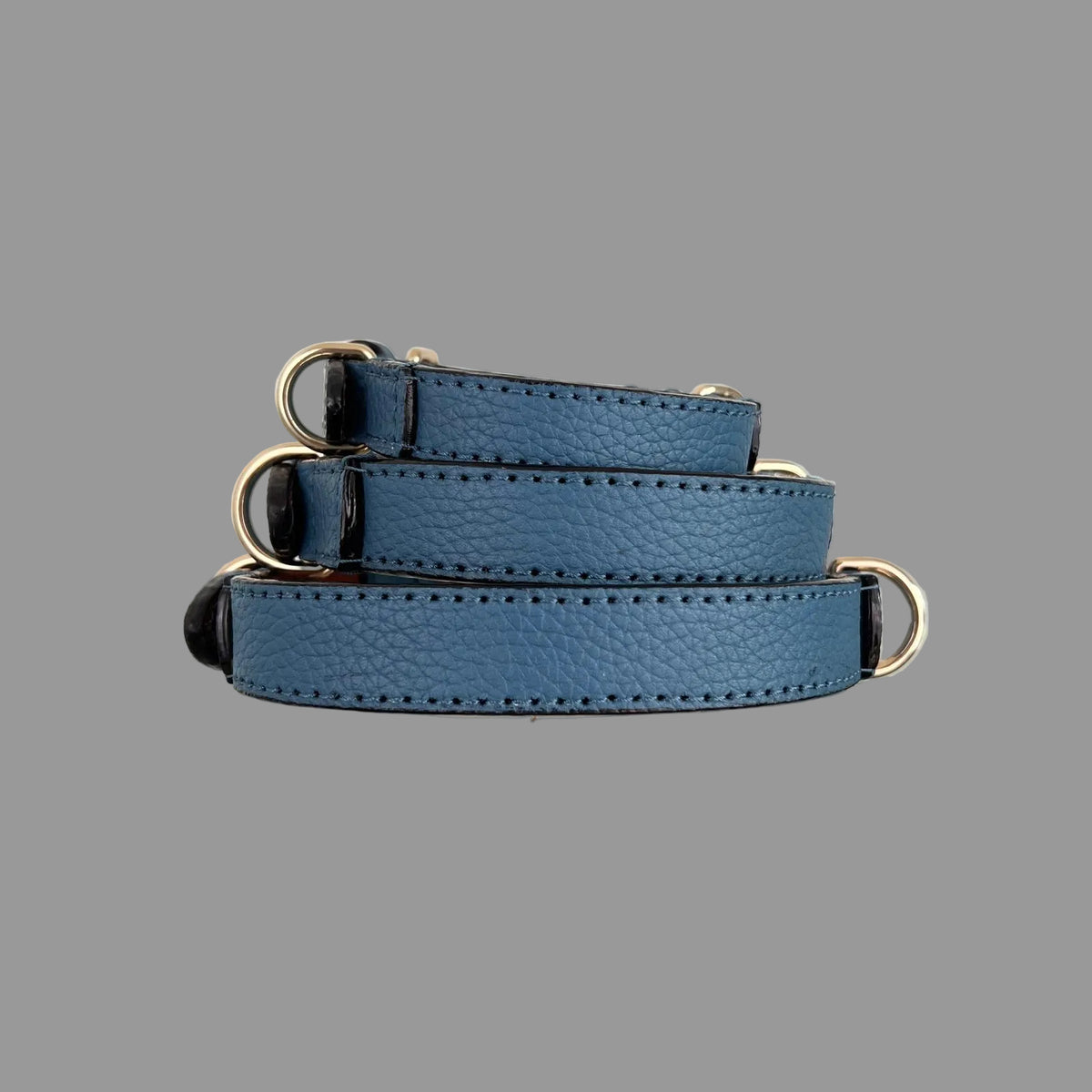 Side view of three denim-blue leather dog collars with pebbled texture, dark edges and gold-tone D-rings.