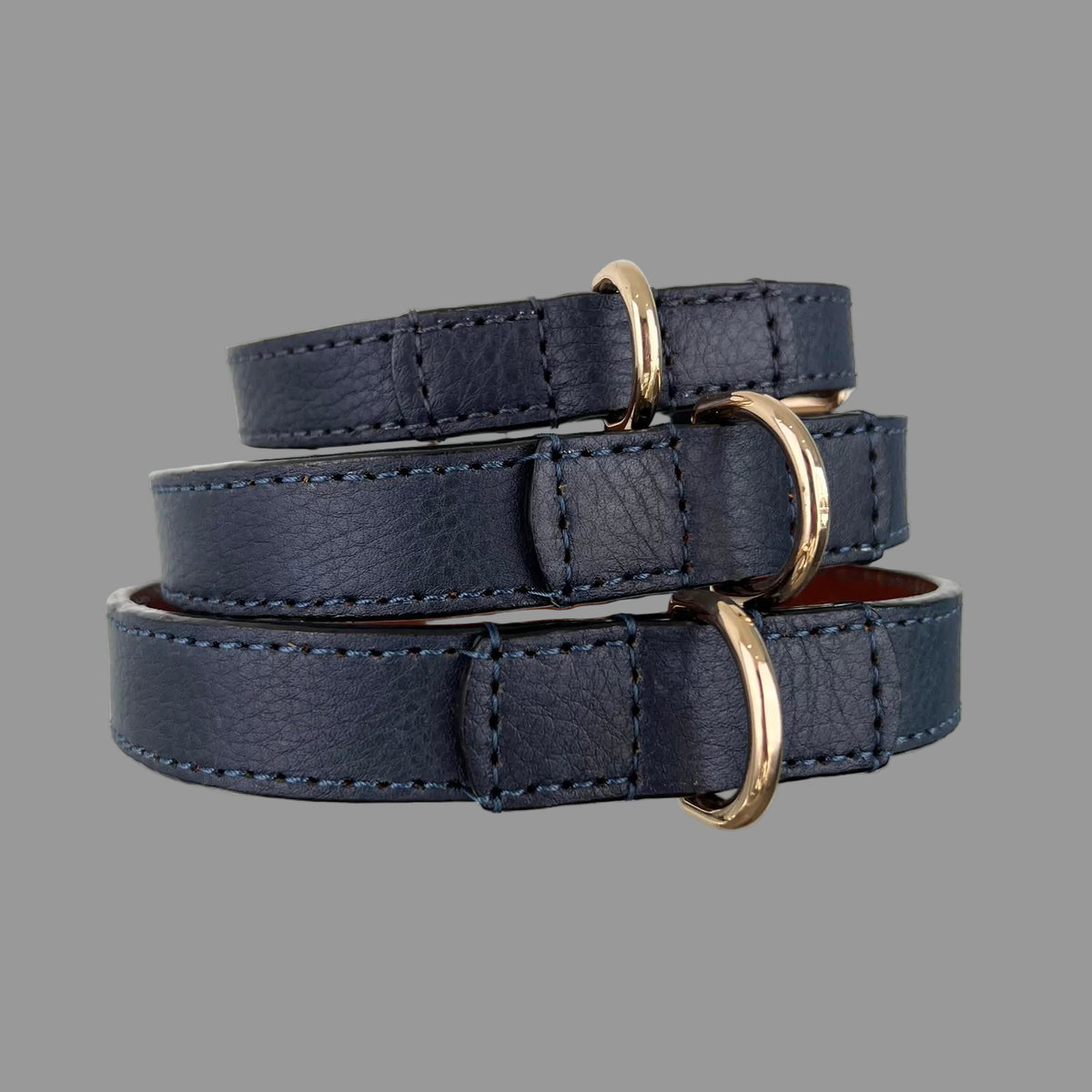 Three navy blue cowhide dog collars showing gold-tone D-rings secured by reinforced leather and blue hand stitching.
